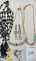 Top view of all jewelry items arranged in tray showing black bead necklace with tassel, gold-tone bar necklace, chandelier earrings, floral blue-stone necklace, and hummingbird brooch with yellow rose earrings.