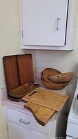 Photo showing two large rectangular wooden trays standing vertically and three wooden bowls stacked next to them, with two wooden cutting boards placed in front on a countertop.