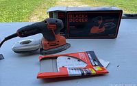 Photo of the Black & Decker Mouse detail sander next to unopened sandpaper package and original box. Shows sander with attached clear dust collection container and detailed grip surface.