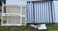 Black metal baby gate, extension, cream plastic baby gate, parts and instructions shown leaning against a table outdoors.