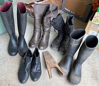 Front view of 5 pairs of different style men's boots: rain boots with orange soles and steel toes, brown winter boots, black quilted winter boots, black leather ankle boots, and tall black rain boots with dirt. Wooden boot shaper also shown.