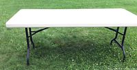 Full view of rectangular Lifetime folding table with white plastic top and black folding legs on grass.