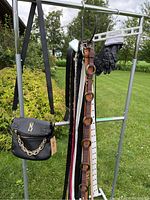Steve Madden black purse with gold chain and MS logo, multiple belts in various colors including white and brown, black leather-like gloves, black Isotoner gloves hanging on metal rack outdoors.