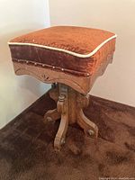 Front and side view of brown upholstered piano stool with carved wooden base showing cushion and decorative nail trim.