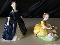 Front view of two Royal Doulton figurines on display including Debbie in dark blue dress and Picnic in yellow dress with picnic setup.