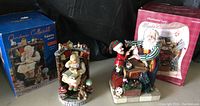 Both collectibles with original boxes shown: Santa Scene figurine seated with book, teddy bear and globe. Santa's Workshop illuminated scene with Santa and elf.