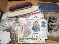 Photo showing various printed sewing patterns for doll clothes, labeled 'Tea Time,' including dresses and accessories, along with fabric-covered press boards and other sewing items.