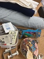 Photo showing three fabric bolts: white, navy blue, and light blue-striped with assorted sewing tools in a clear blue zippered pouch and a plastic container with buttons and snaps. Also visible is a decorative round tin and a metal clamp part.
