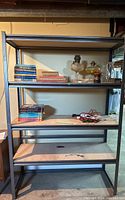 Full view of sturdy metal shelf with four particle board shelves, on a concrete basement floor. Visible wear and some items present on shelves but not included.