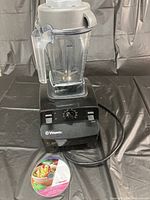Vitamix Aspire blender base with clear blending container on top and power cord coiled to side. Instructional DVD placed in front of blender on black surface.
