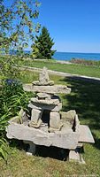 Front angle photo showing the concrete bench with Inukshuk sculpture outdoors, near grass and a pathway with water in the background.