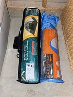 Two camping tents packed in carry bags on a concrete floor. The left tent is in a green-yellow bag labeled 'Dome Tent'. The right tent is in a blue and orange bag labeled 'Eagle's Camp'.