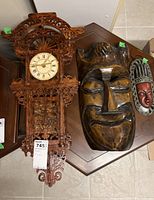 Photo showing carved wooden wall clock alongside two ethnic carved wall masks on a table.