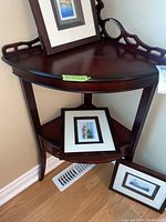 Overview of the two-tier corner display table with framed pictures placed on it (not included in sale). Shows the top and middle shelves and partial legs.