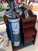 Photo showing golf bag filled with assorted golf clubs next to a wooden organizer stand with multiple open shelves, placed on a carpeted floor.