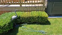 Wide view showing four metal plant hooks with curved tops and a metal heron statue placed outdoors on grass near a hedge and deck.