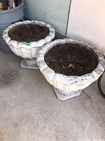 Pair of white outdoor planter urns filled with soil, showing weathered condition and dirt marks on their surfaces, photographed on a concrete floor indoors.