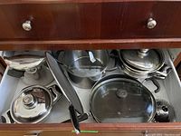 Drawer open showing assorted stainless steel pots and pans with lids and handles inside a cabinet.