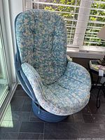 Full view of blue painted rattan chair with large, floral-patterned cushion covering seat and back, positioned near window.