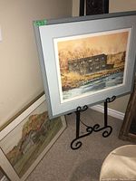 Framed signed and numbered print by Gordon Peters displayed on black metal easel, showing artwork of rustic wooden building near river in autumn colors.