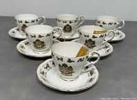 Six white bone china teacups with matching saucers, each cup featuring a different zodiac sign crest and gold trim, arranged on grey surface.