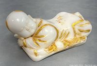Front and side view of hand carved white stone child figurine reclining on a pillow, displaying gold accents mostly on robe details and hair.