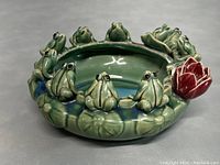 Angle view of green glazed lotus-shaped bowl with multiple sculpted frogs around the rim and a red lotus flower accent.
