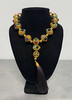 Front view of the 16 inch copper necklace displayed on a black jewelry bust, showing gold-tone textured beads with multicolored stone accents and a dark brown tassel.