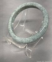 Close-up photo of the natural jade bangle on a clear display stand showing its pale green marbled color and smooth polish.