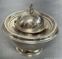 Front angle of round, footed sterling silver dripper with hinged lid closed and finial knob visible