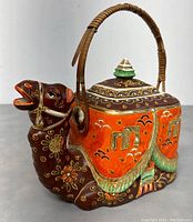 Front and side view of the camel-shaped ceramic teapot showing moriage decoration, floral patterns, and wicker handle.