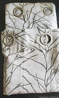 Two folded curtain panels showing silver metal grommets and silver/gray fabric with green branch design.