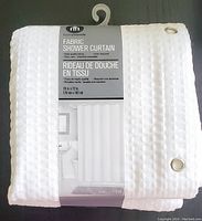 White folded fabric shower curtain with waffle texture edging and metal grommets, still on original packaging with Hometrends label visible showing size and care instructions.
