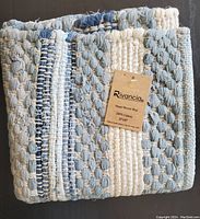 Folded view of the hand woven Rivancia cotton rug showing thick weave with blue, grey, and cream colors and attached brand tag.