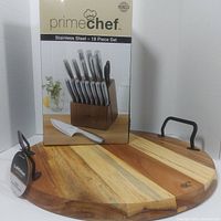 Photo of the Prime Chef 18-piece stainless steel cutting tool set box alongside the round acacia wood serving board with metal handles.