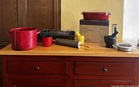 Overview of assorted kitchen items including red speckled pots and mug, roasting pan with rack, rolling pin, measuring cups, mortar and pestle, strainer, and boxed red pot.