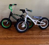 Two children's balance bikes side by side on a wood floor, one black and green, one white.
