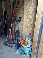 Shot showing long handled garden tools leaning against shed wall including shovels, rakes, hoes, and a turquoise bucket with garden stakes.