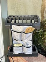 Front view of black plastic outdoor storage organizer with multiple compartments and attached fabric organizer holding broom and brushes.