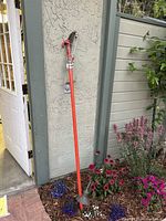 Full view of long handled Procut extended pruner leaning against a wall in a garden setting with flowers below, showing orange handle and cutting head at top.
