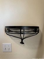 Front view of black semi-circular metal wall shelf mounted on wall showing decorative circular elements in frame and curled support at bottom.