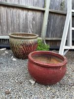 Two vintage drip paint glazed planter pots placed on gravel ground outside against a wooden fence; one pot solid red, the other with multicolor drip glaze