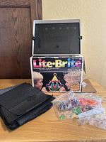 Full view of the Lite-Brite pegboard with multiple black screens stacked next to it, plastic bags filled with colored pegs, and the original box behind it.