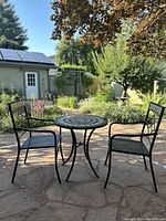 Full view of the patio set showing round mosaic tile table and two black metal mesh chairs in outdoor garden setting