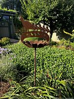 Rust-colored metal silhouette yard ornament of a pheasant and smaller birds on a curved band with flower-shaped metal bowl, staked in garden among plants and greenery.