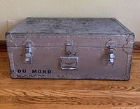 Closed metal footlocker on floor showing front with metal latches and handle and stenciled text.