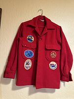 Front view of red wool jacket with five embroidered scouting patches sewn on, hanging on a hanger against a beige wall.