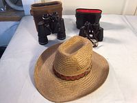 Front view of the straw cowboy hat with colorful band and two pairs of binoculars with cases behind it.