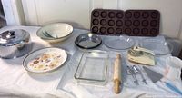 Full view of collection laid out on white tablecloth including baking pans of various types, mixer with beaters, rolling pin, serving accessories, and glass food cover.