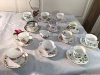 Various bone china teacups and saucers displayed on white cloth, showing individual floral designs with gold accents, including teacups paired with matching saucers.